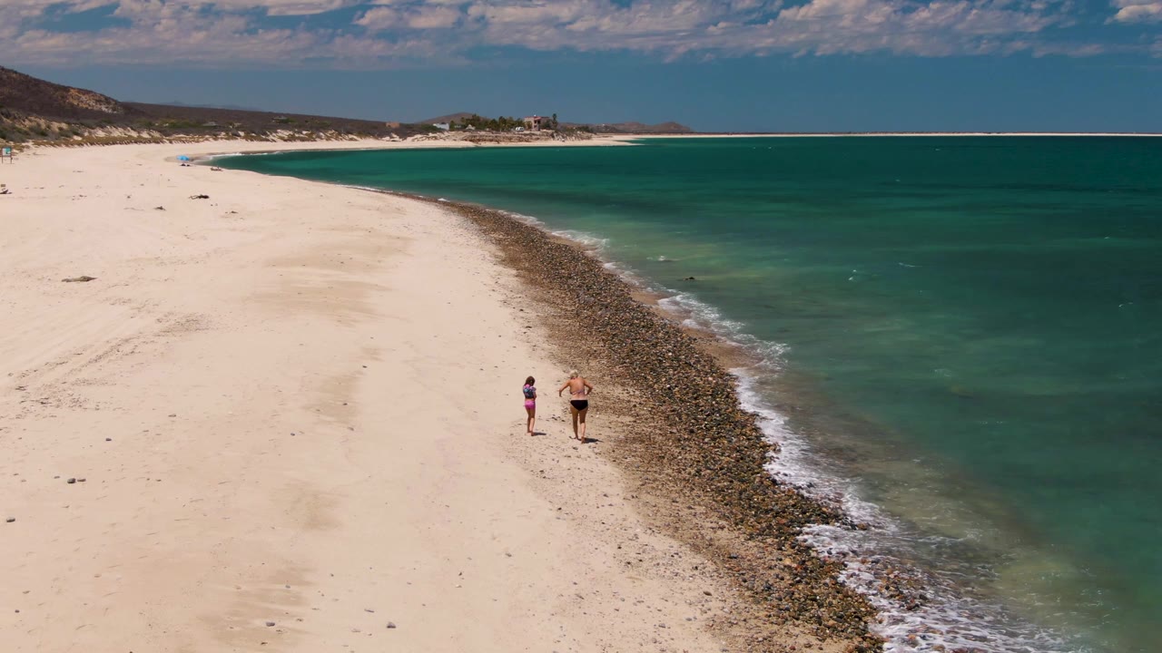 Adult and child walking along pale sand near the water's edge.