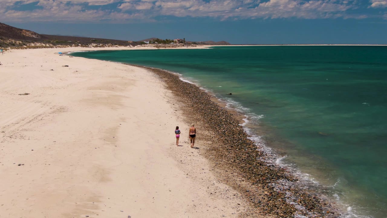 At 0:02 on Barakas Beach, the two walkers appear slightly farther along the shoreline while the curved rocks and thin wave line separate sand from turquoise ocean.