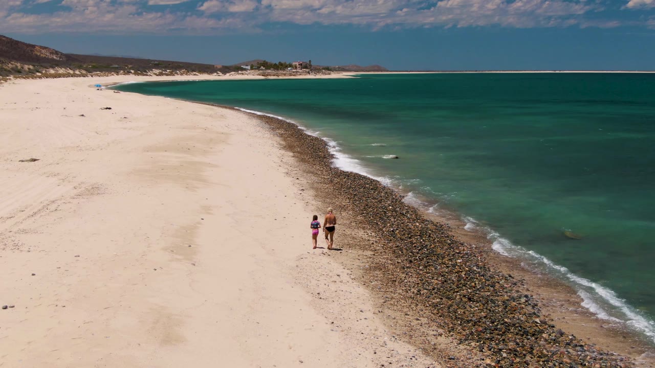 At Barakas Beach in Baja California Sur, two people walk together on pale sand beside a diagonal rocky edge and turquoise ocean under scattered clouds.