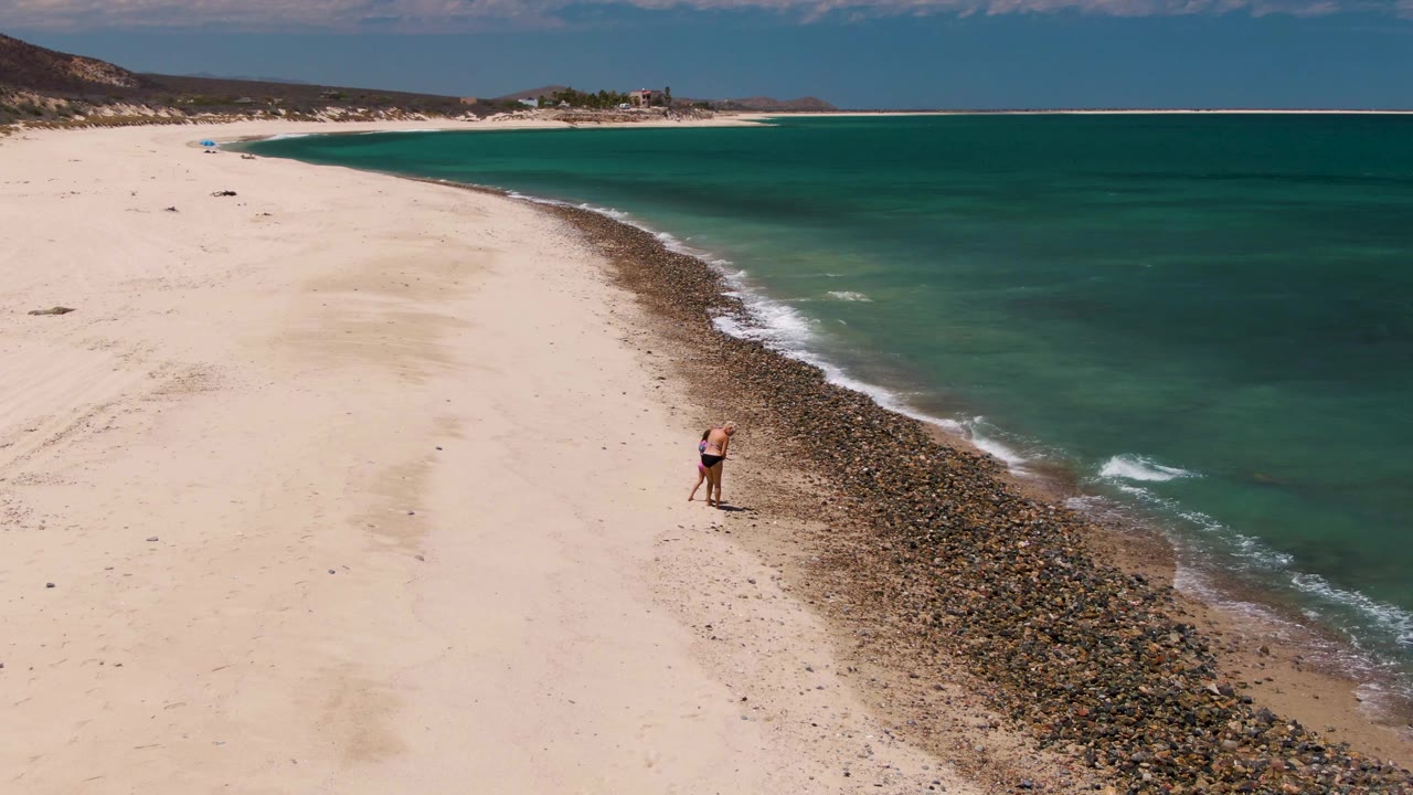 At Barakas Beach in Baja California Sur, one visible person remains near the rocky strip while turquoise water and a broad sweep of empty sand fill the frame.
