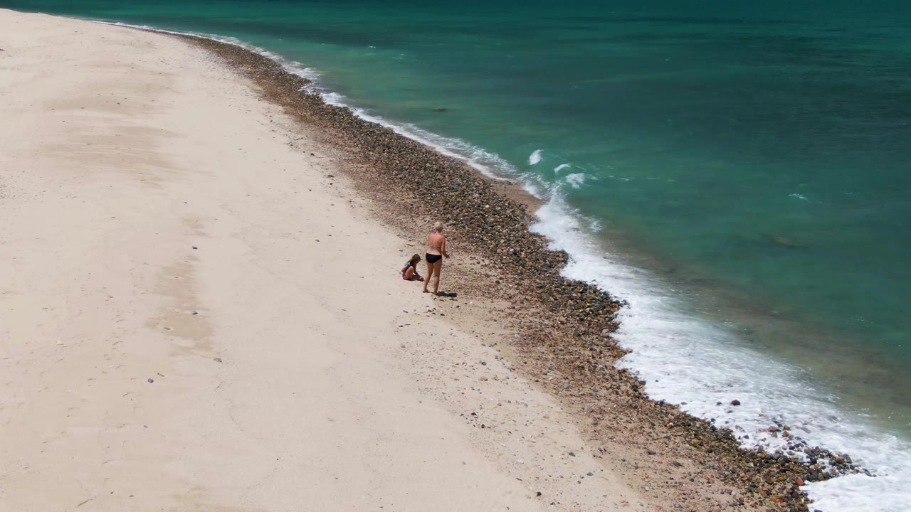 At Barakas Beach in Baja California Sur, a person bends near the edge of a rocky shoreline as turquoise water breaks into foam beside pale sand.