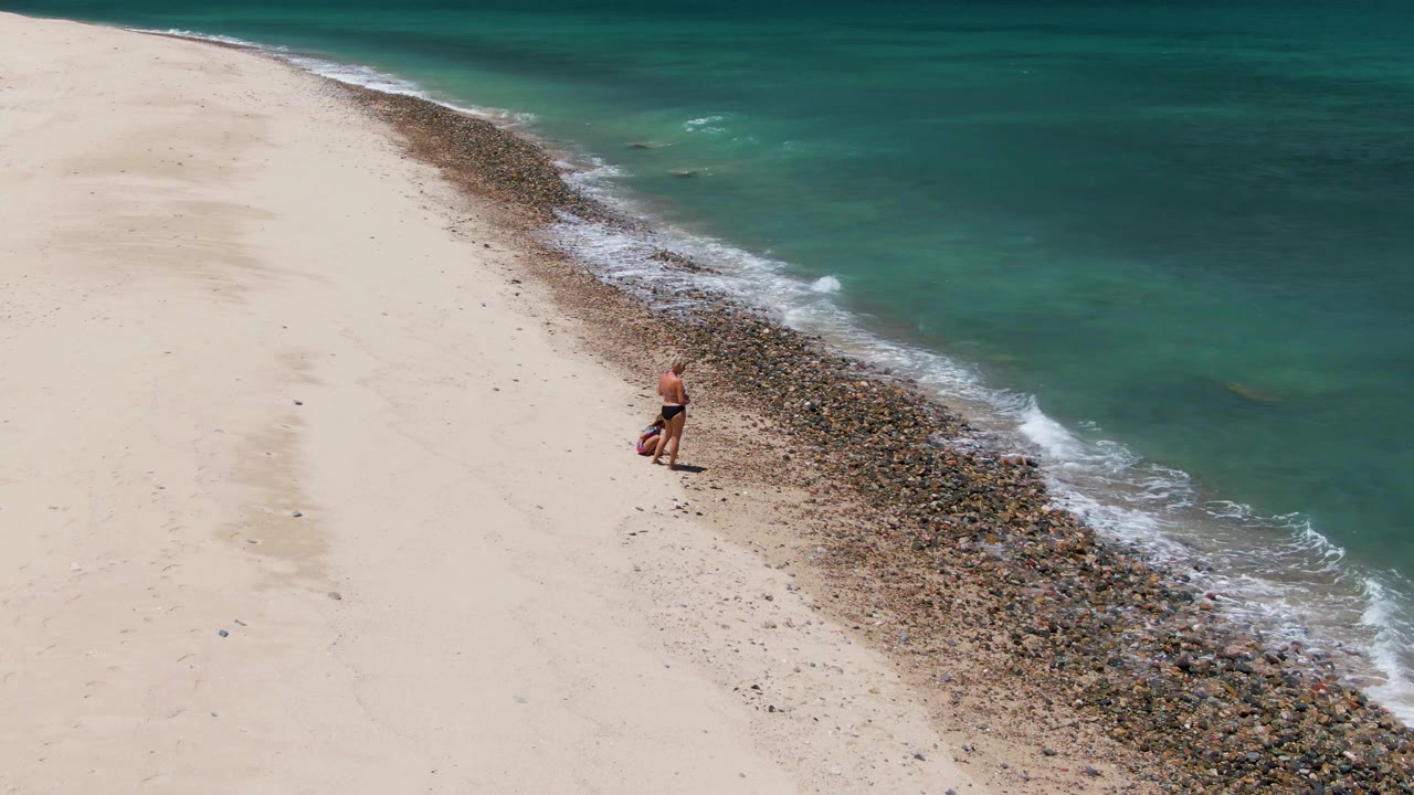 At Barakas Beach in Baja California Sur, an aerial frame shows an adult near a dark rocky surf line with pale sand on the left and turquoise water on the right.