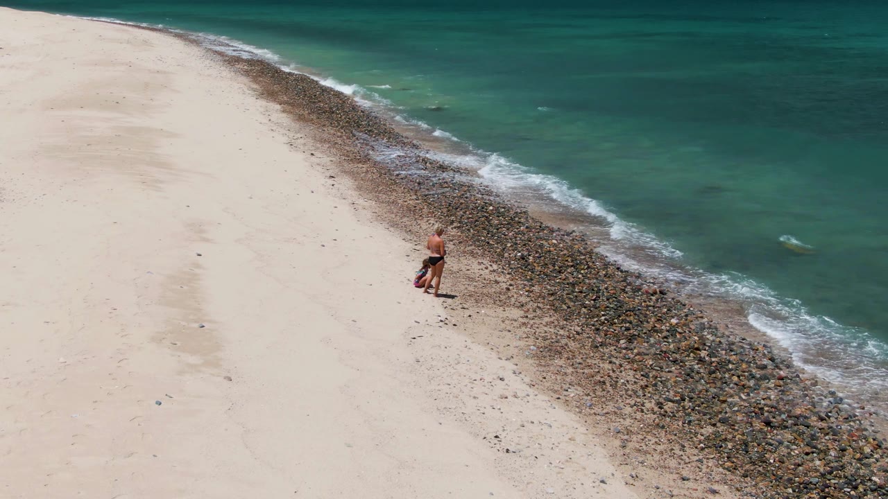 At 0:02, the aerial view shows the adult slightly farther along the curve of rocks, with pale sand stretching left and green-blue water rippling to the right at Barakas Beach.