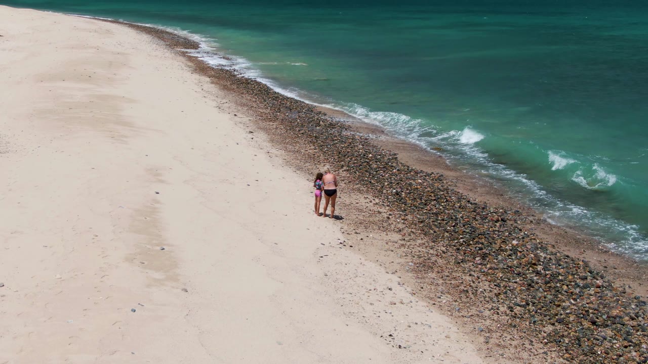 Two people stand on pale sand at Barakas Beach in Baja California Sur, Mexico, beside a diagonal strip of pebbles and turquoise surf at the start of the clip.