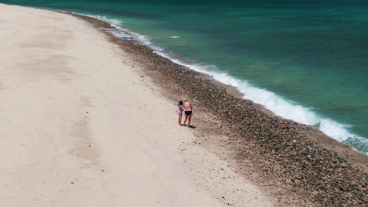 Two people on pale sand near a pebbly shoreline and clear water.