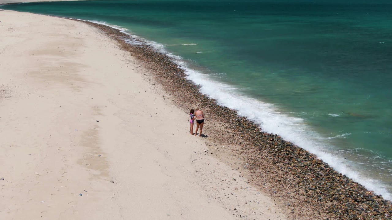 An overhead view at Barakas Beach shows the two people slightly right of center, with white surf spreading across the pebbly band between sand and turquoise ocean.