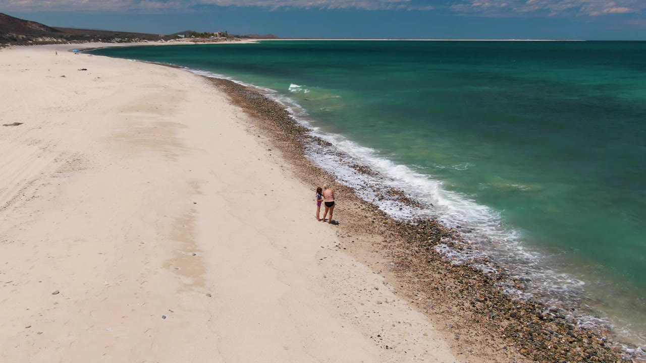 Aerial frame from Barakas Beach, Baja California Sur, showing two distant people walking near the rocky edge of the turquoise ocean with wide white sand to the left.