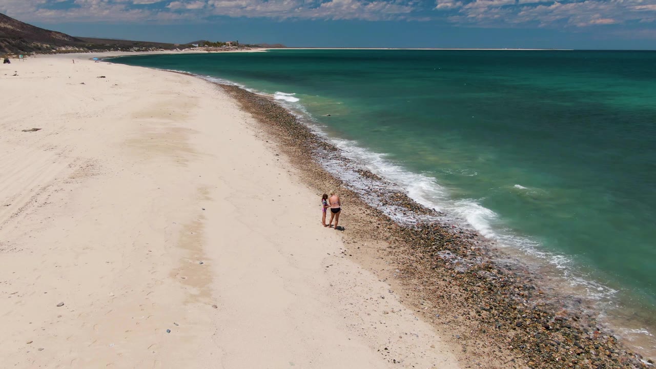 Two people walk along the rocky edge of a sandy beach beside turquoise water.