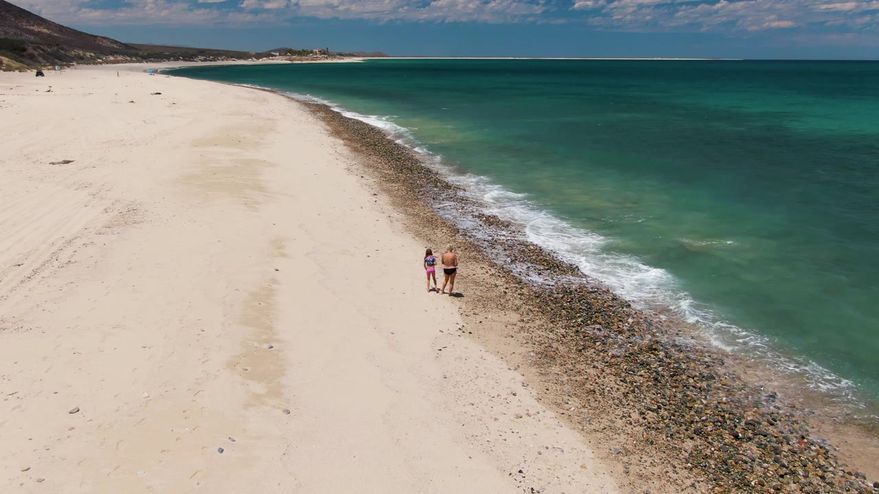 Barakas Beach aerial still showing the couple near the right side of the sand, with gentle wave foam touching the rocks and a broad empty beach stretching toward distant hills.