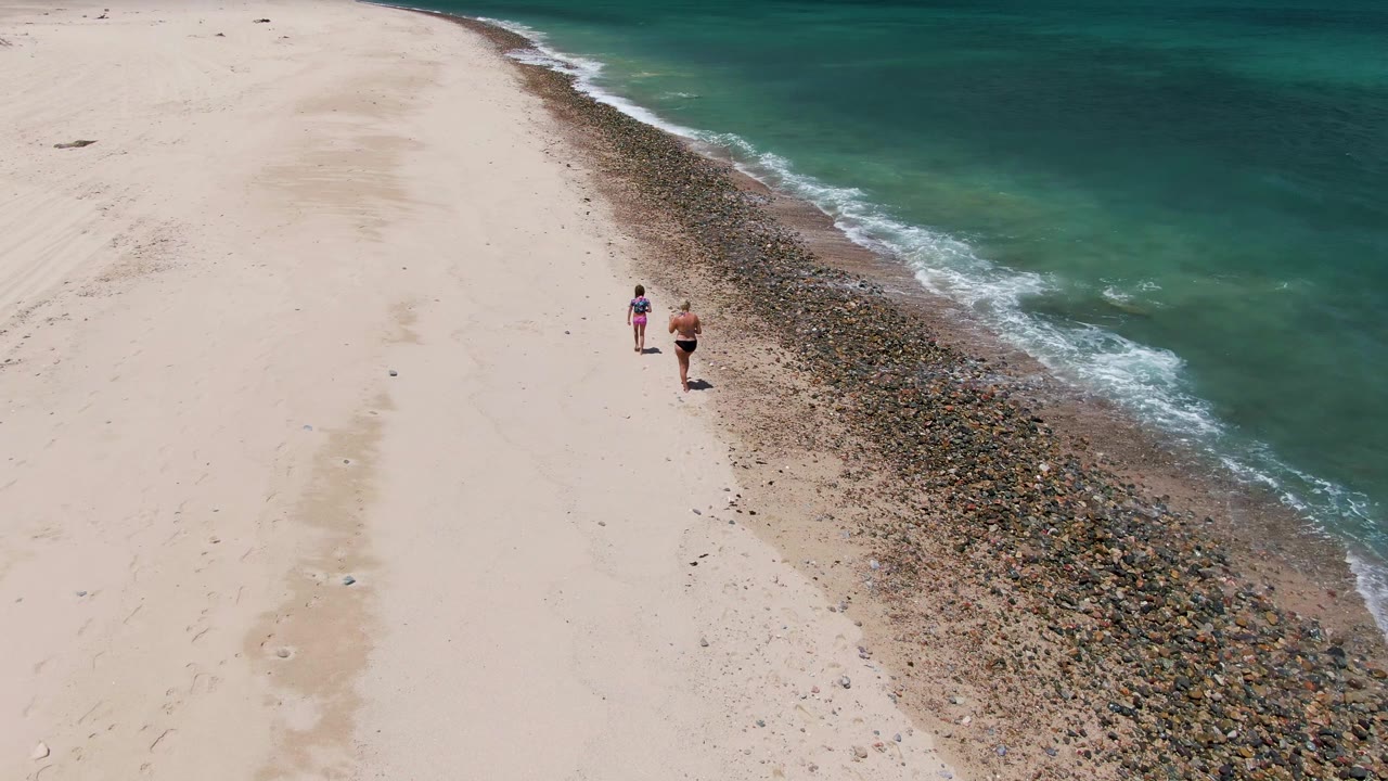 Two people walk along the pale sand at Barakas Beach in Baja California Sur, with a dark pebble strip and turquoise ocean water along the right side.