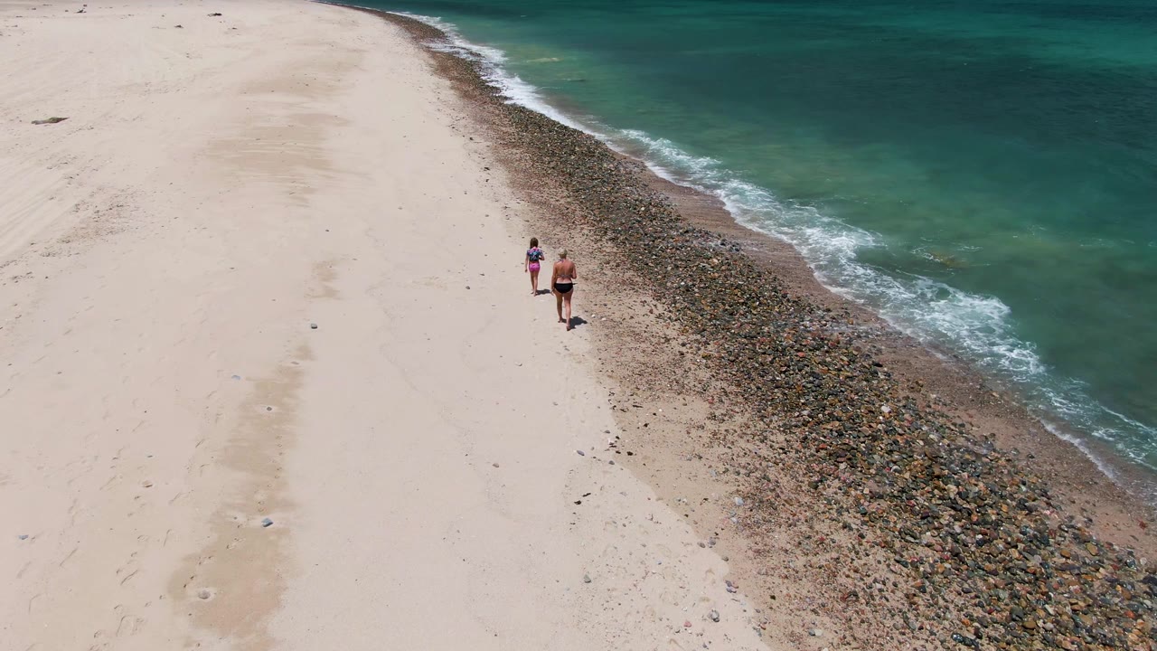 Two people continue walking near the pebble line on a beach.
