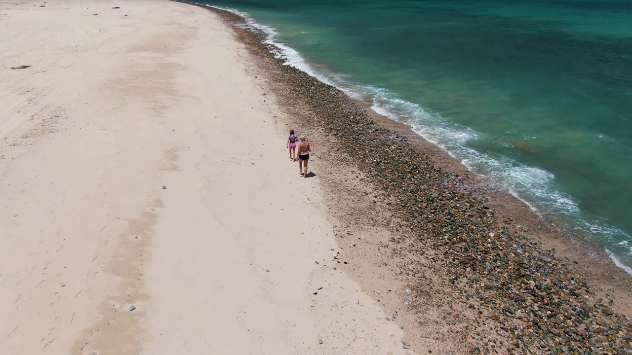 Two people walk near the upper center of the aerial frame at Barakas Beach, with broad pale sand to the left and clear turquoise water to the right.