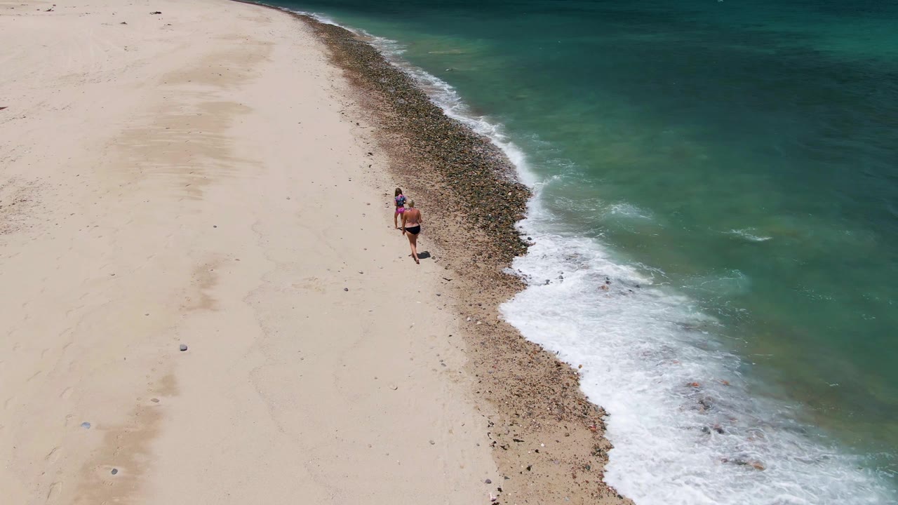 A woman walks near the waterline on Barakas Beach in Baja California Sur, with pale sand on the left and turquoise surf breaking along a dark shoreline band.