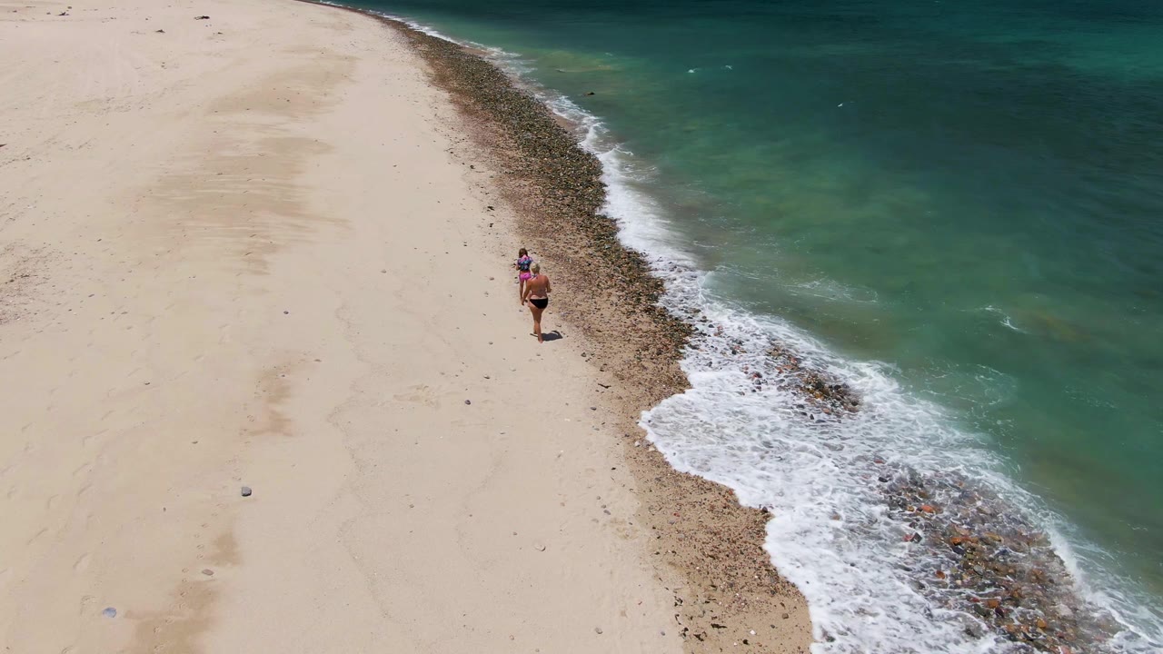 Aerial view of a woman walking beside foamy surf on sand.