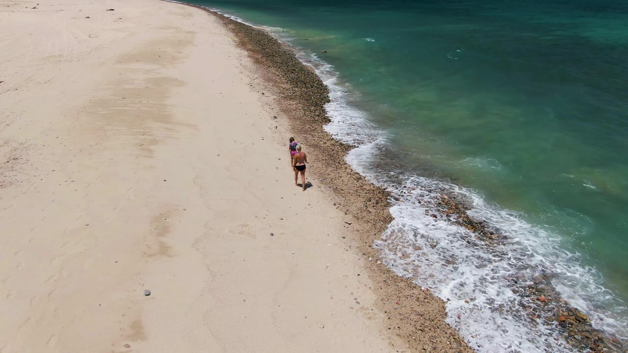 At Barakas Beach, the walker appears farther along the diagonal shore while white water curls around the darker band between dry sand and turquoise ocean.