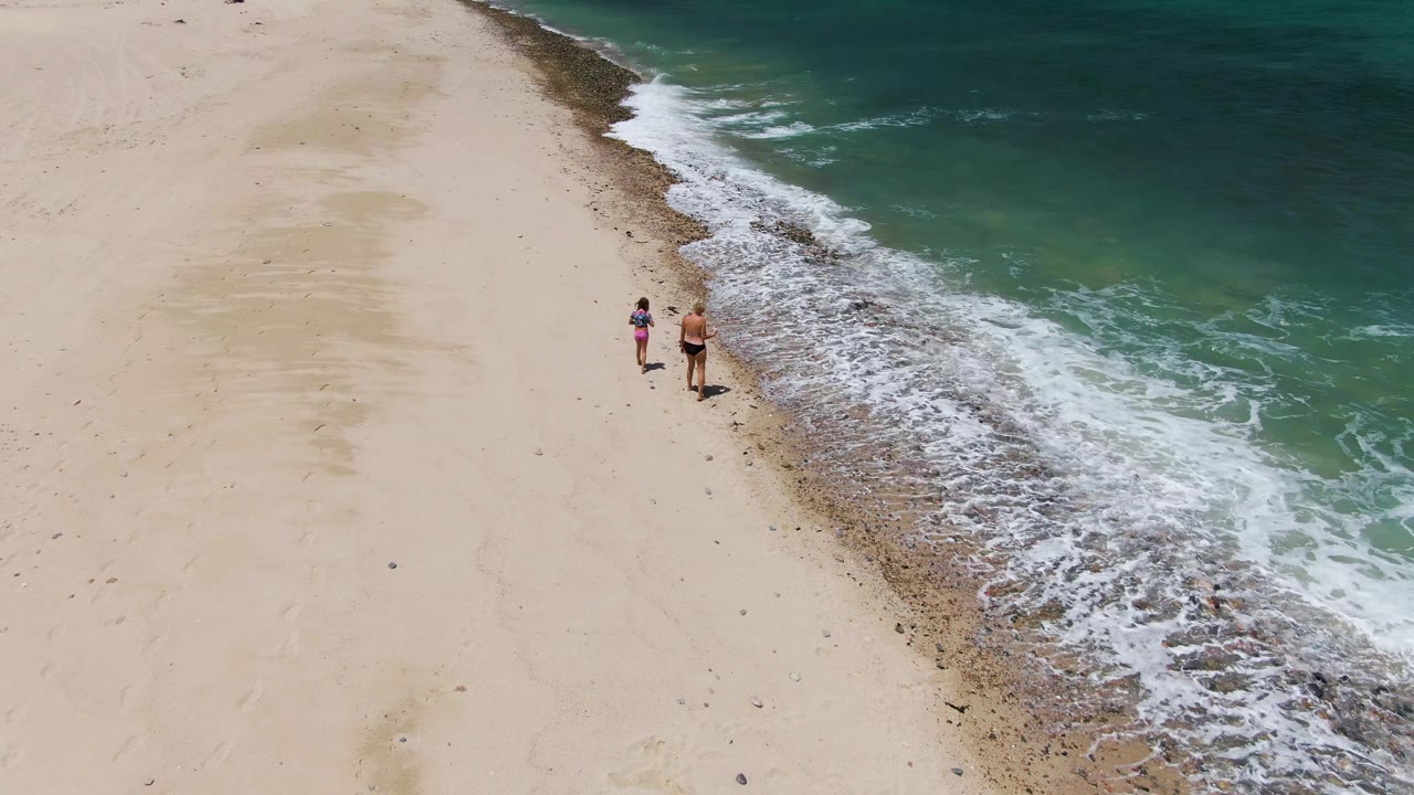 Two people walk near the shoreline at Barakas Beach, Baja California Sur, with pale sand on the left and green-blue ocean water on the right.