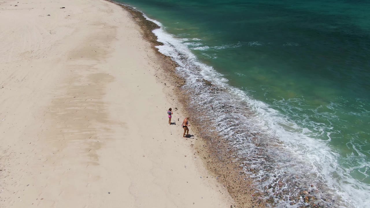 Two walkers move along the shoreline as waves foam beside them.