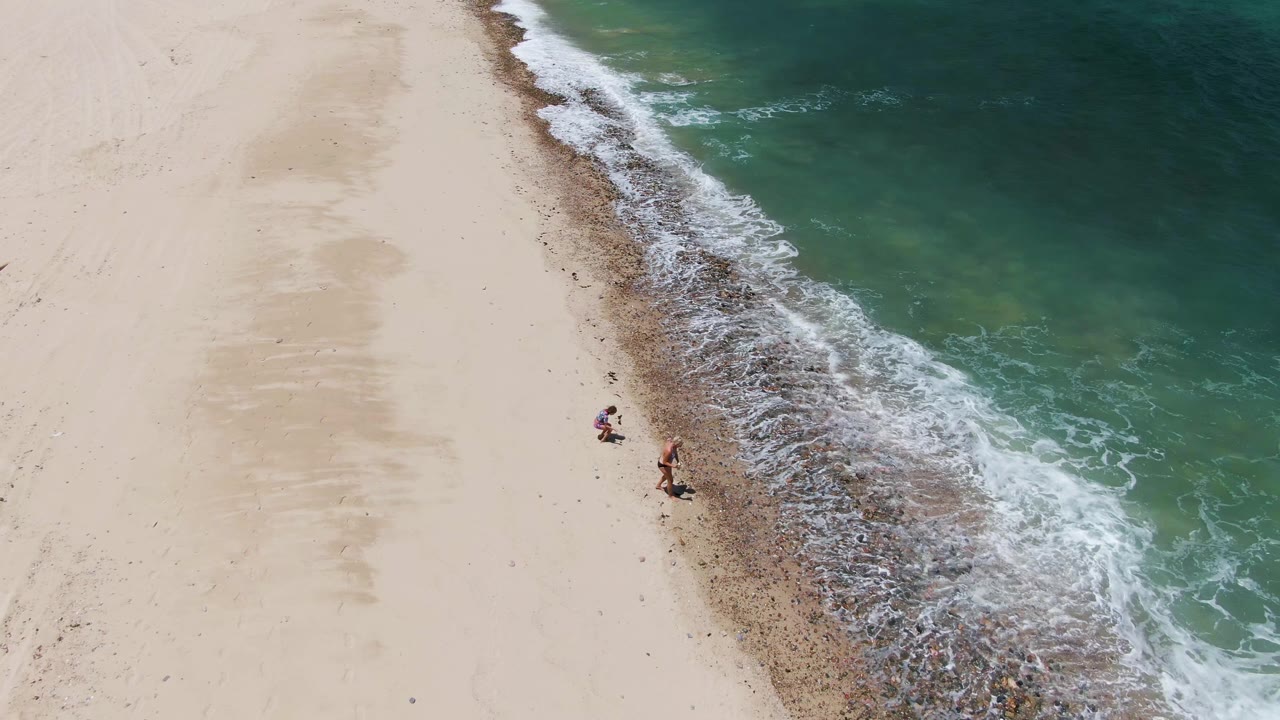 The walkers appear farther along the Barakas Beach shoreline, with a wider band of white foam and green-blue water filling the right side of the frame.