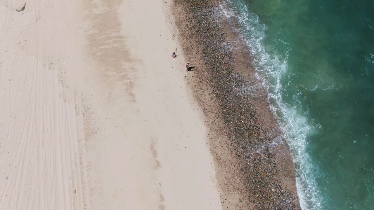 Overhead view of Barakas Beach with white sand on the left, turquoise surf on the right, a dark wet shoreline band, and small people visible near the upper sand area.