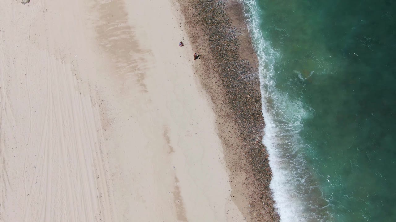 Aerial shoreline view with pale sand, green-blue water, and foam along the edge.