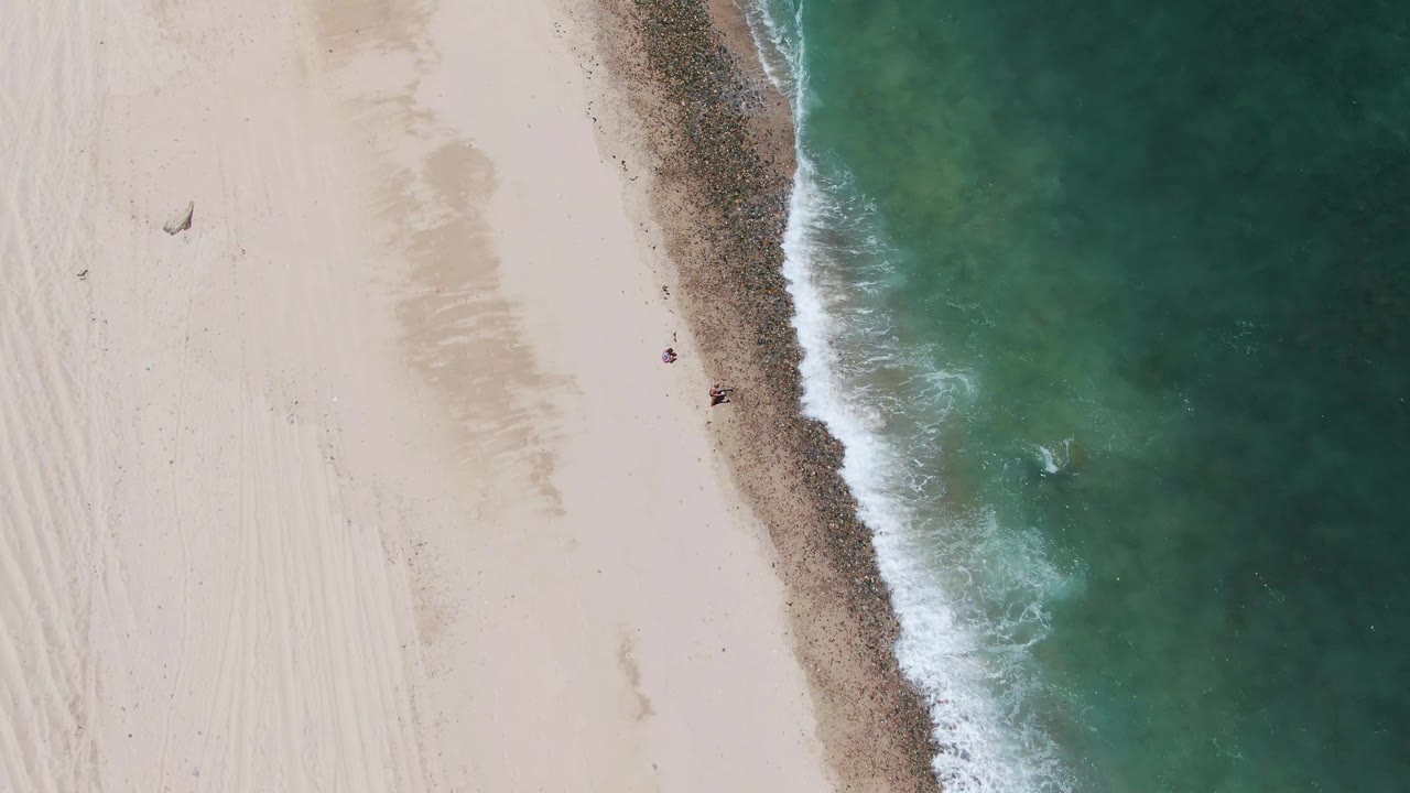 Top-down view of Barakas Beach with broad white sand, turquoise water, a foamy shore edge, and small people visible near the upper and mid shoreline areas.
