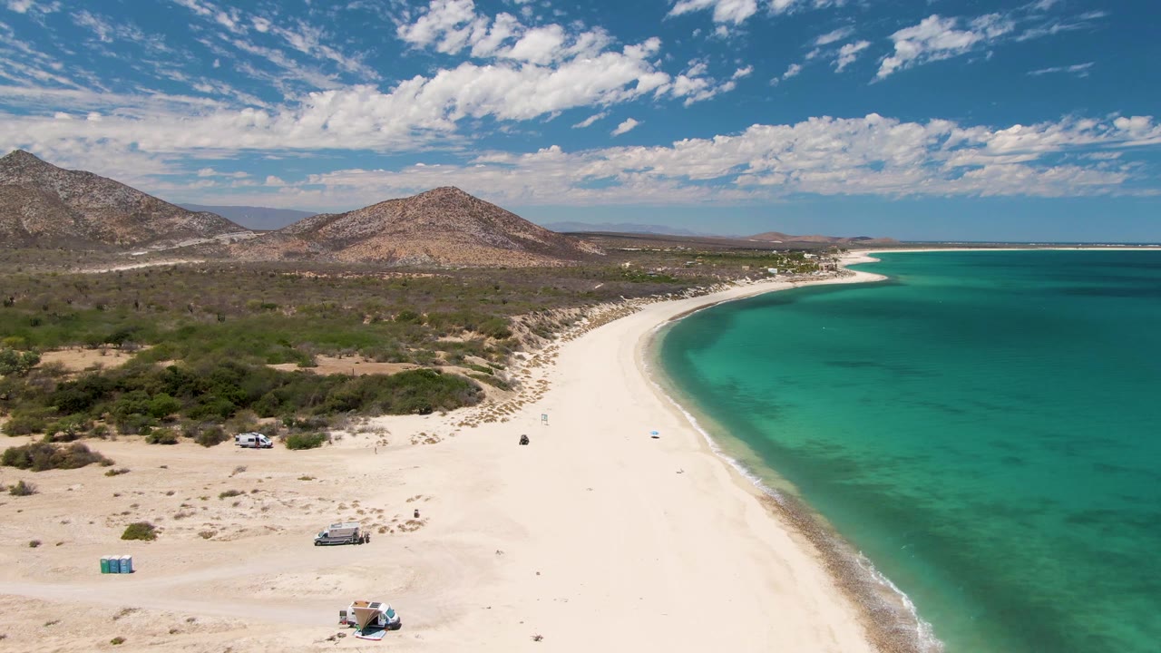 Opening aerial frame over Barakas Beach, Baja California Sur, with white sand curving along turquoise water, scrubland inland, low mountains beyond, and small vehicles and people visible on the beach.
