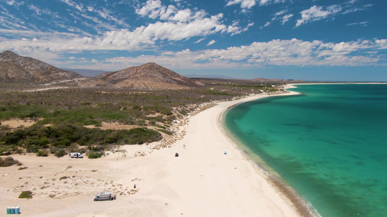 Barakas Beach appears in a high oblique aerial view with the pale sand arc leading toward the horizon, green-blue water along the right edge, inland scrub, mountains, and tiny beachgoers and vehicles below.
