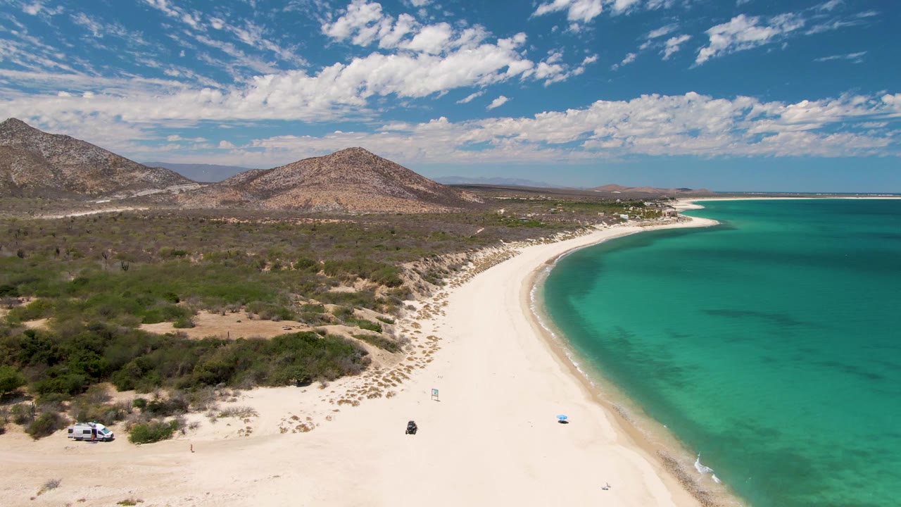 Aerial frame at Barakas Beach, Baja California Sur, showing curved white sand beside turquoise water, dry hills, scrub vegetation, a white vehicle near the lower left beach edge, and a tiny distant person-like figure on the sand.