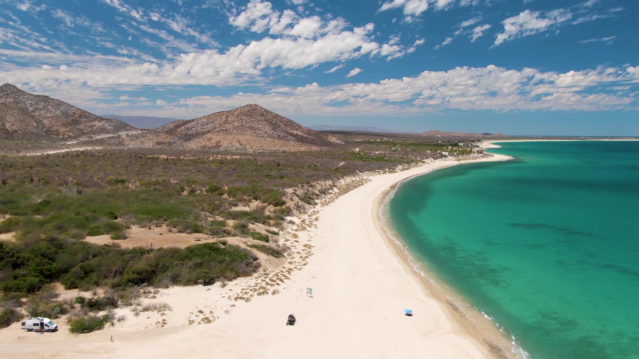 Aerial frame at Barakas Beach, Baja California Sur, with the white vehicle near the left edge, a tiny distant person-like figure farther down the sand, turquoise water to the right, and dry hills behind the shoreline.
