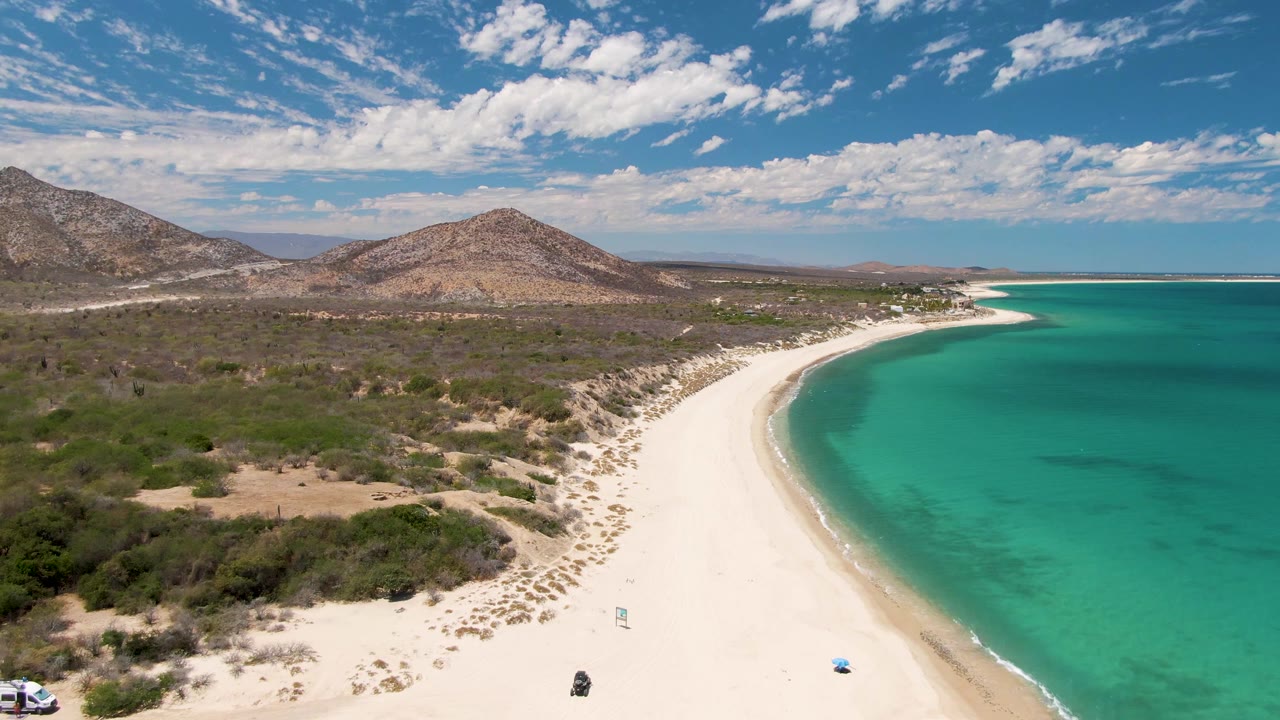 Aerial frame at Barakas Beach, Baja California Sur, showing the shoreline curve more centered, the turquoise ocean filling the right side, dry scrub and hills inland, a white vehicle clipped at the far left edge, and no recognizable people.