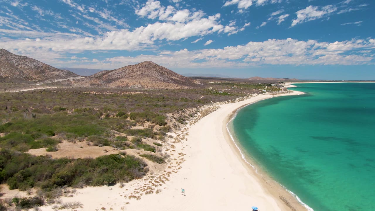 Aerial frame at Barakas Beach, Baja California Sur, with the beach curve running from lower center toward the distant shore, clear turquoise water on the right, scrubby desert vegetation on the left, hills in back, and no visible people.