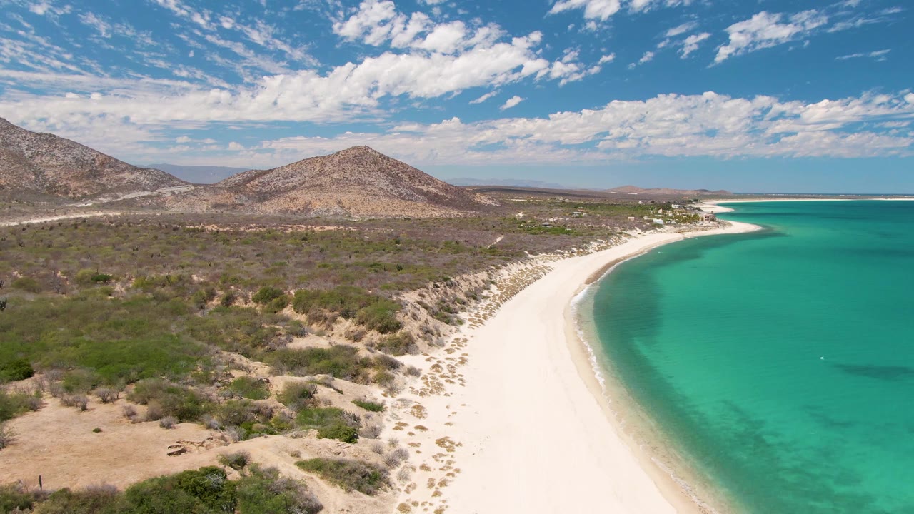 Drone-style view of Barakas Beach with turquoise water on the right, a pale sandy shoreline through the center, scrubby terrain on the left, distant hills, and no visible people.