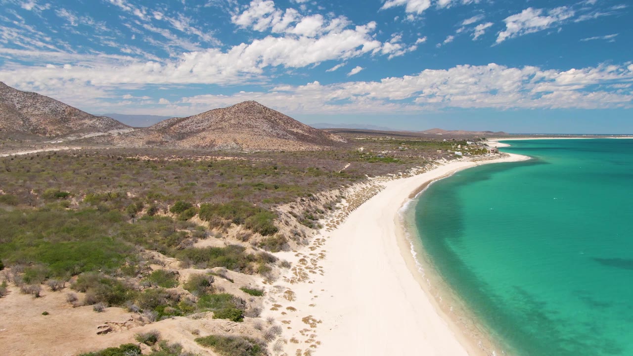 High aerial view at Barakas Beach showing the white shoreline sweeping toward the distance, bright turquoise shallows, low desert vegetation, brown hills, scattered cloud cover, and no visible people.