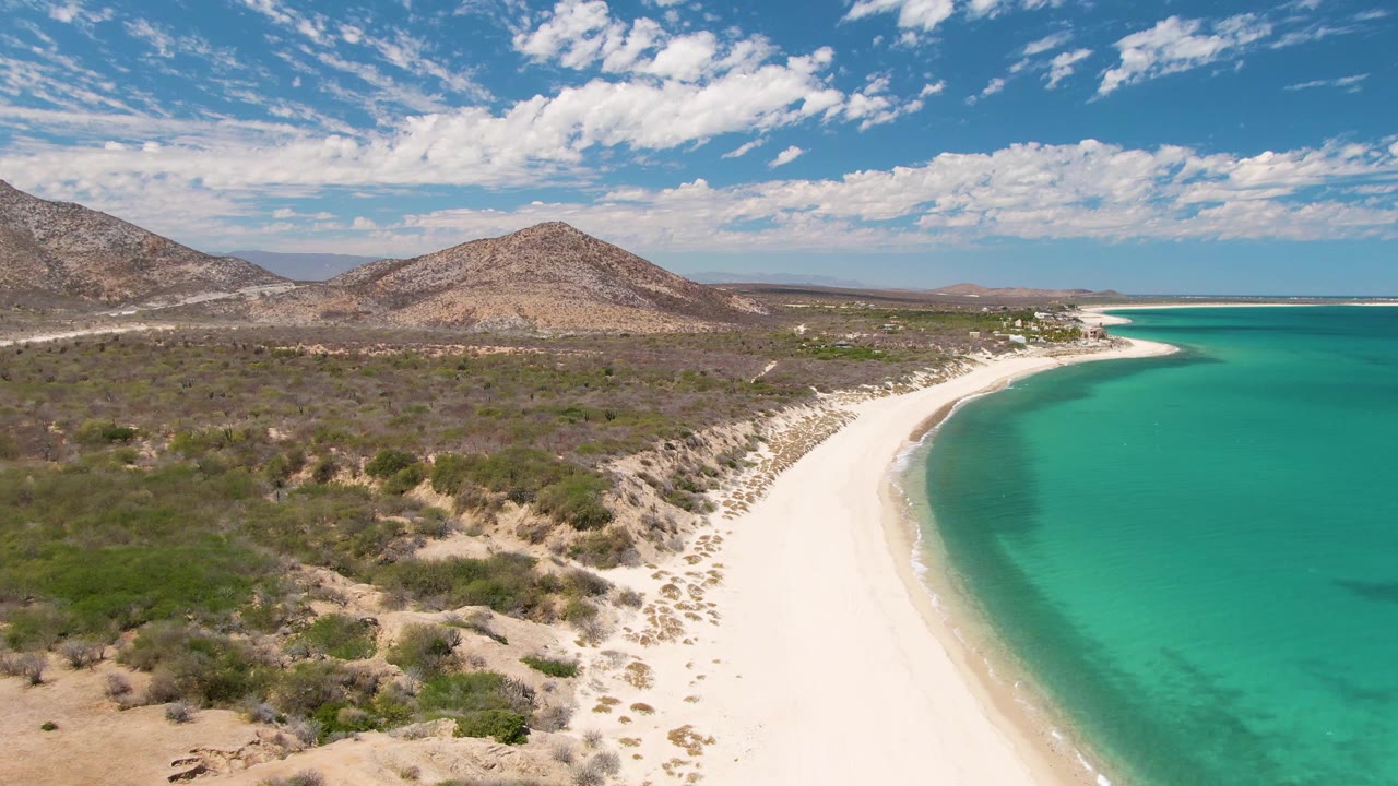 Wide aerial frame of Barakas Beach with the ocean occupying the right side, the white sand arc running through the middle, dry brushland to the left, low hills on the horizon, and no visible people.