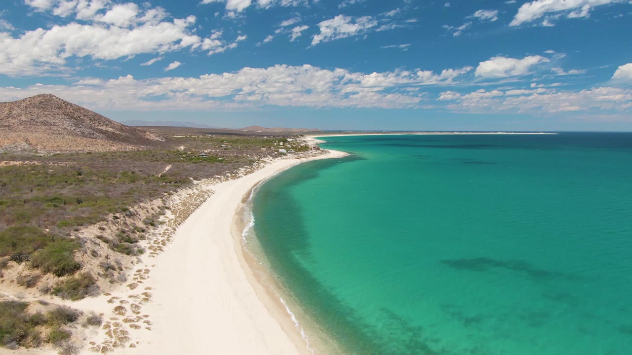 Opening aerial frame at Barakas Beach, Baja California Sur, showing a white sand curve, turquoise shallows, low scrubland, a brown hill, scattered clouds, and no visible people.