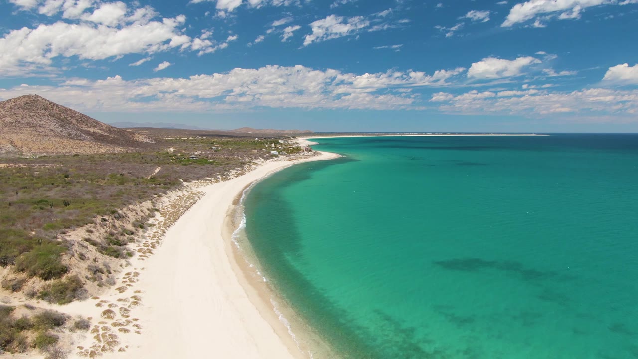 One second into the Barakas Beach aerial, the shoreline arc remains centered with pale sand on the left, deeper turquoise water on the right, sparse vegetation inland, and no visible people.