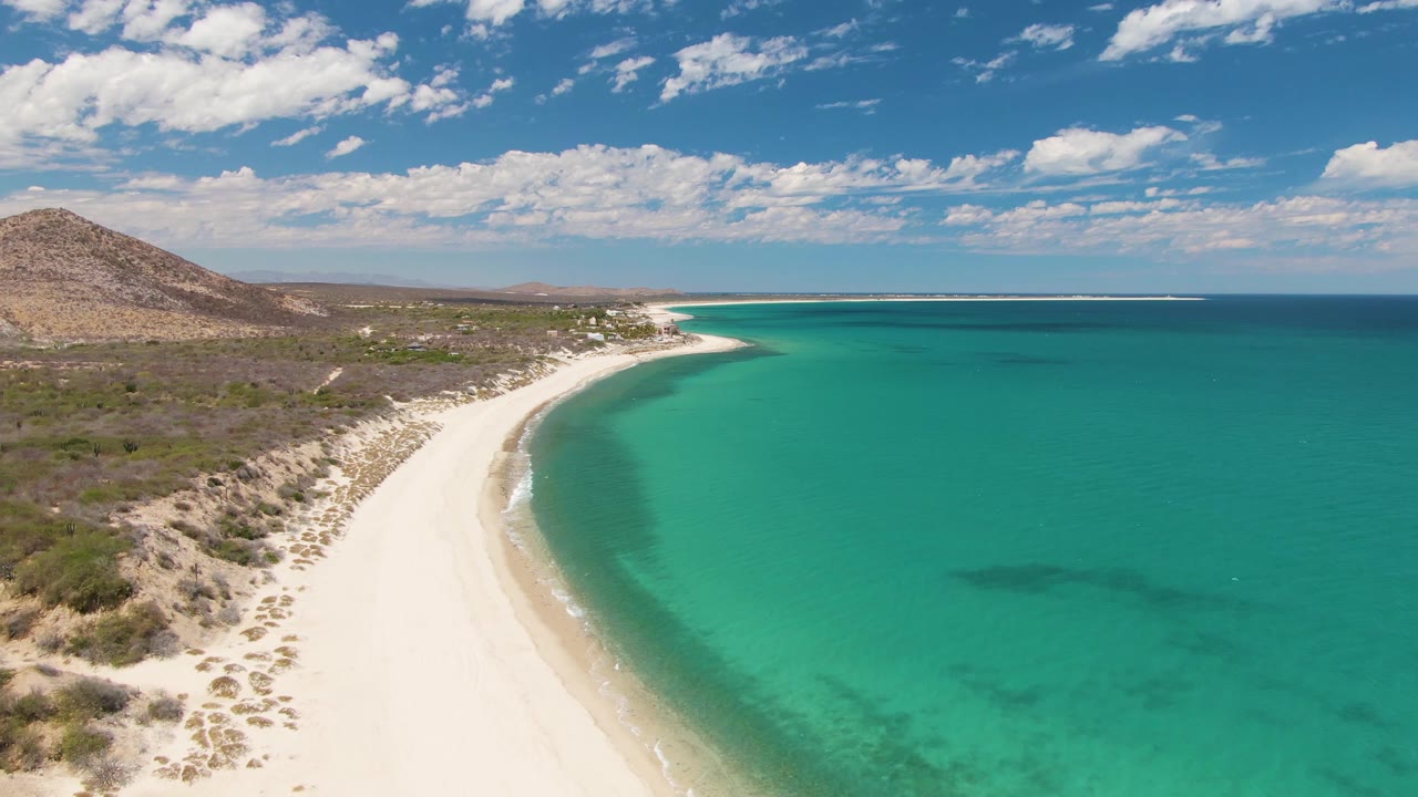 At 0:02, the aerial view looks along the Barakas Beach shoreline as small waves trace the sand, turquoise water fills the right side, inland scrub and a low hill sit to the left, and no visible people appear.