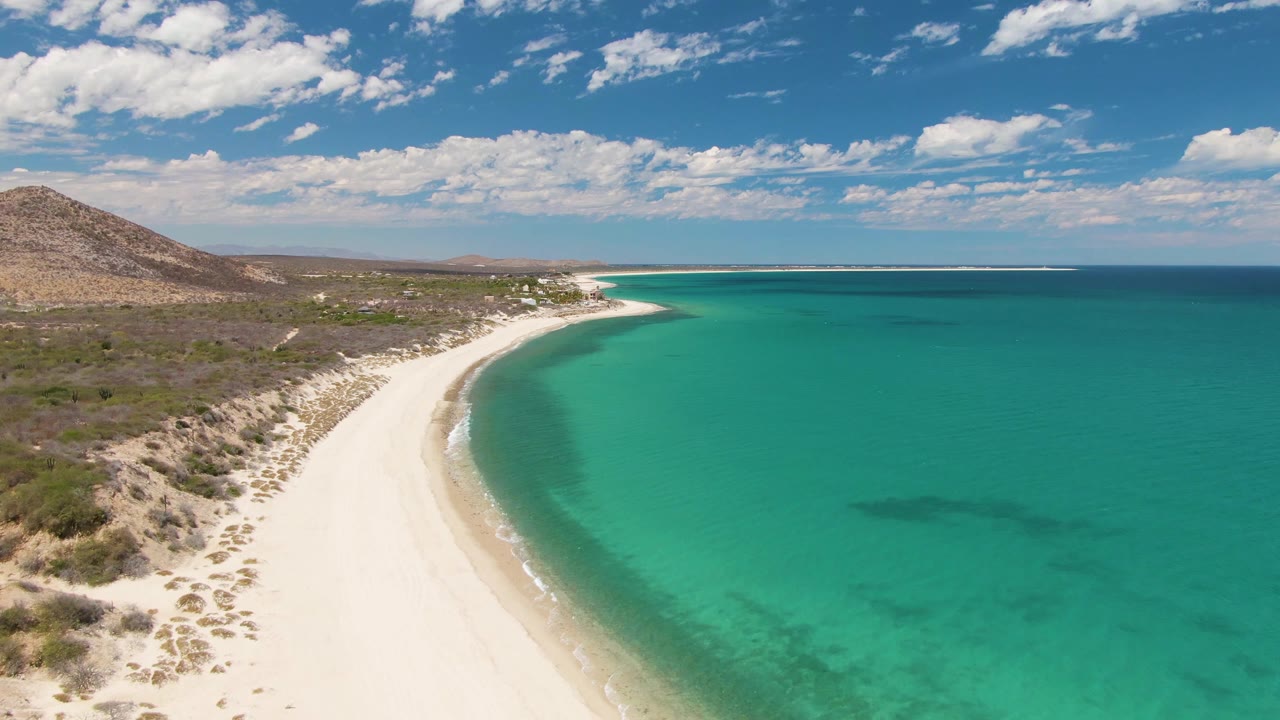 Aerial beach view with white sand foreground, turquoise sea, hill, and clouded sky.