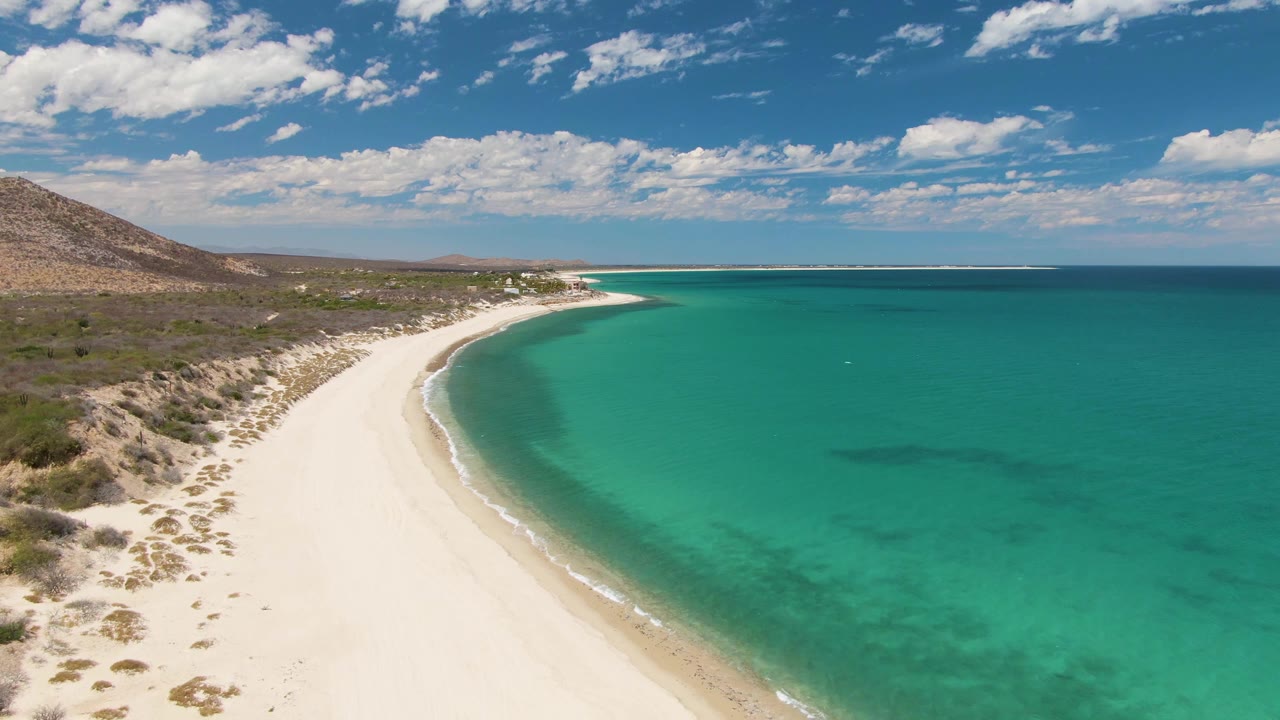 At 0:01, Barakas Beach fills the left side of the frame while green-blue water spreads to the right, with a thin surf line, inland scrub, distant hills, and no visible people.