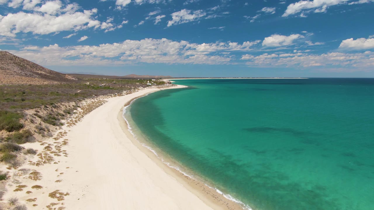 At 0:02, the aerial view shows Barakas Beach with pale sand sweeping toward the distance, darker patches under the turquoise water, scrubby dunes, inland hills, and no visible people.