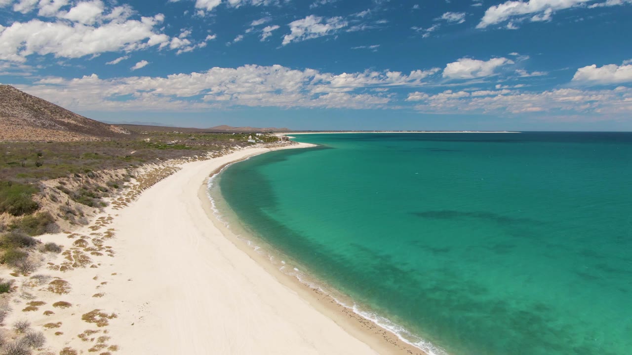 At 0:03, Barakas Beach appears as a wide diagonal strip of white sand with turquoise ocean on the right, low vegetation on the left, hillside terrain beyond, and no visible people.