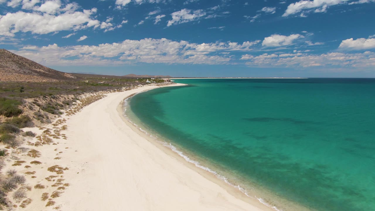 Opening aerial view of Barakas Beach in Baja California Sur, with white sand curving along turquoise water, scrub-covered land at left, a brown hillside beyond, and no visible people.