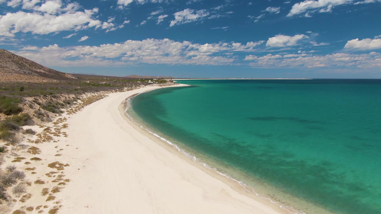 Aerial frame of Barakas Beach showing the shoreline arc slightly farther along the waterline, with pale sand in the lower frame, turquoise ocean to the right, hillside to the left, and no visible people.