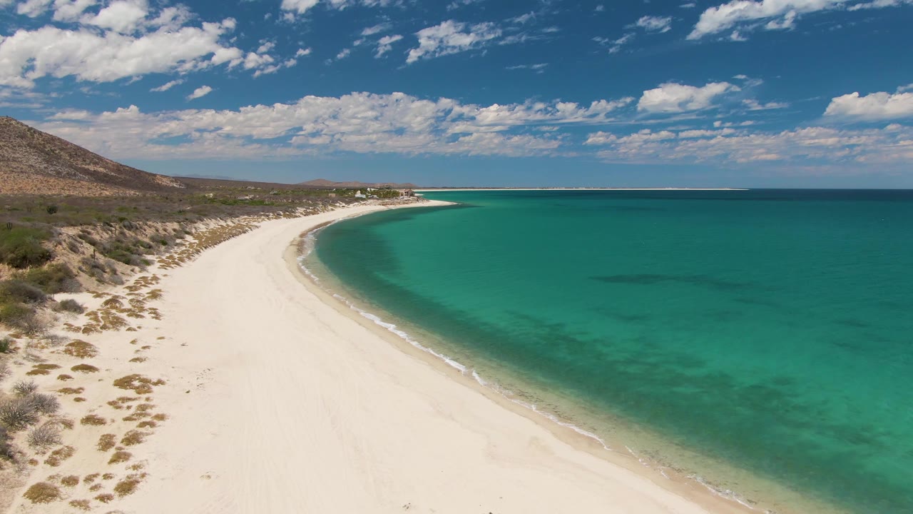 Barakas Beach from above at two seconds, with the sand curve leading toward a distant point, deeper turquoise water on the right, dry coastal vegetation on the left, and no visible people.