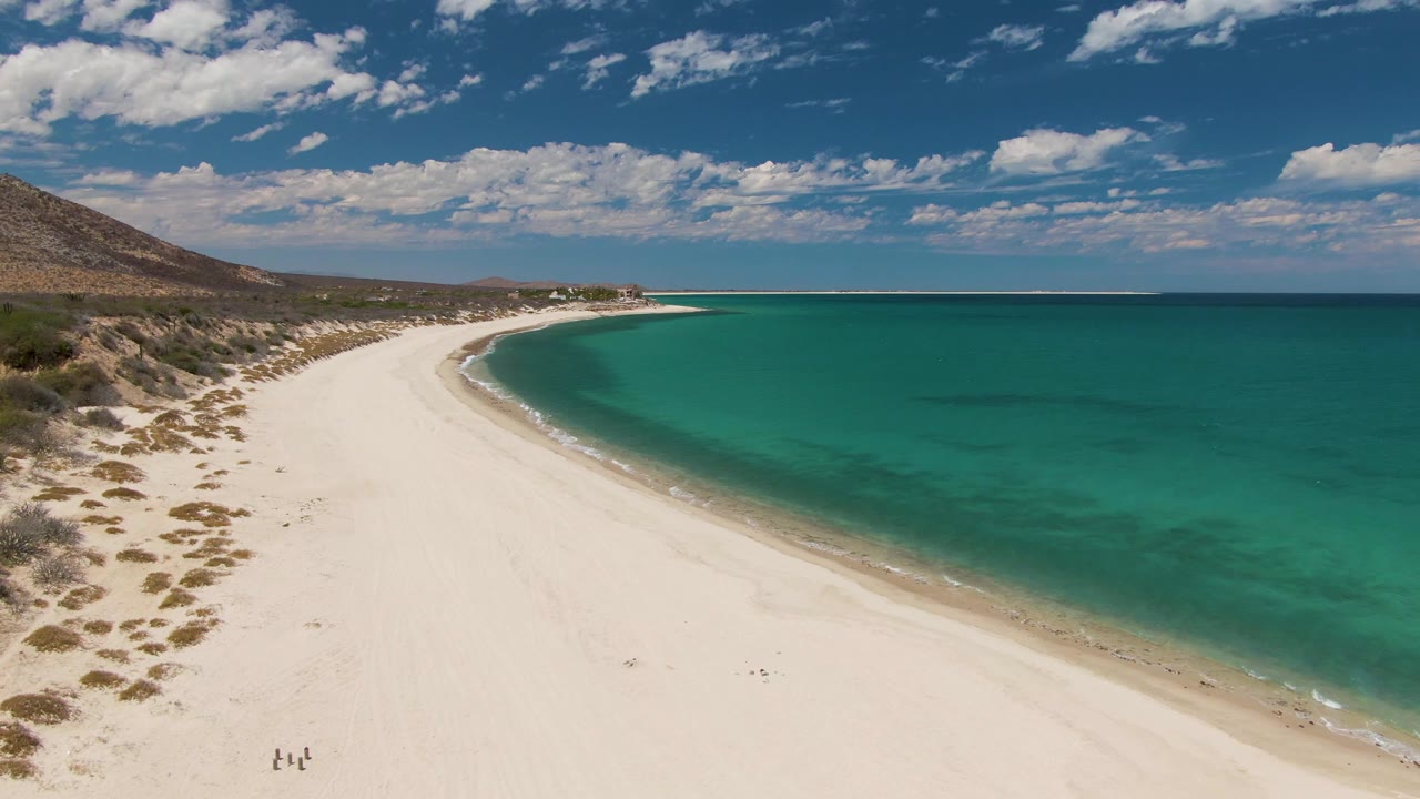 A high aerial view at Barakas Beach, Baja California Sur shows a broad white sand curve, turquoise ocean on the right, scrubland and hillside on the left, and tiny people on the lower beach.