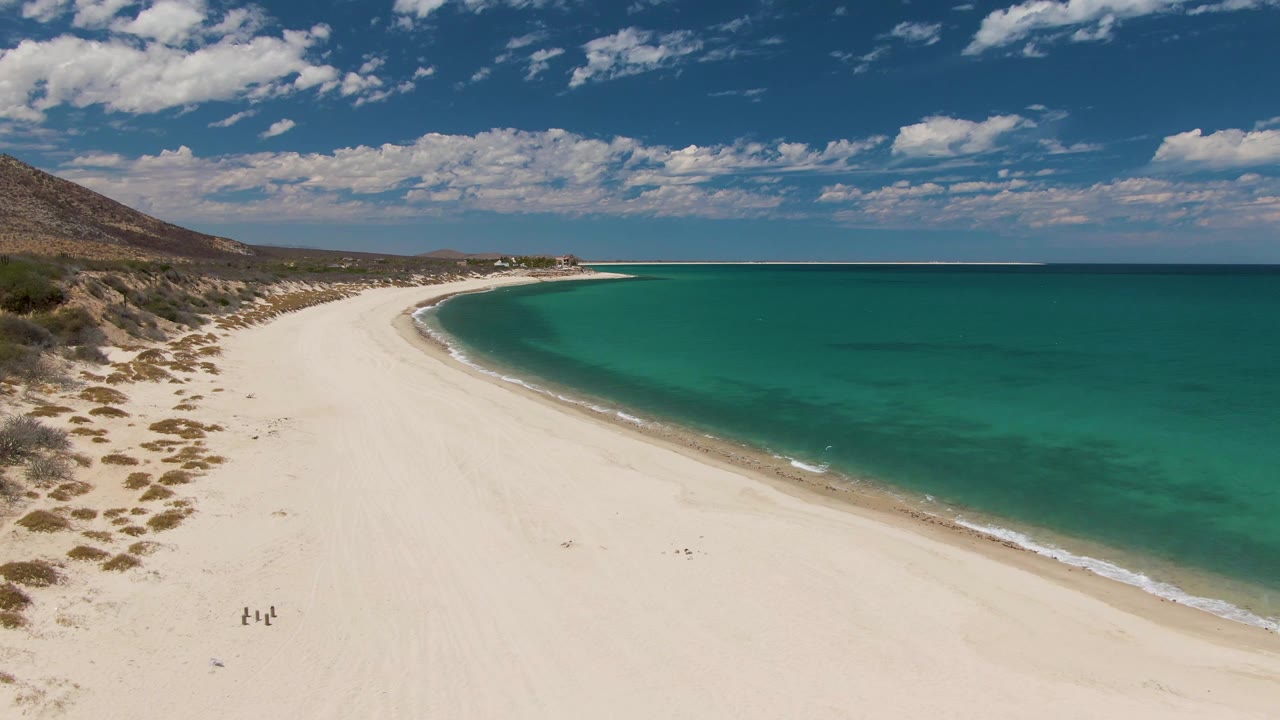 At 0:02, the aerial view shows the same Barakas Beach shoreline with walkers farther into the lower sand area, a thin band of waves, open turquoise water, and a distant low horizon.
