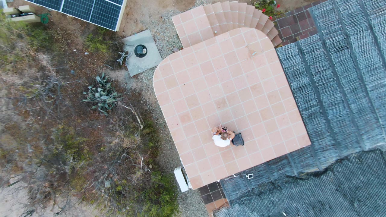 Overhead drone frame in Cabo Pulmo, Baja California Sur showing a pale patio roof, nearby solar panels, desert vegetation, and one person visible on the patio.