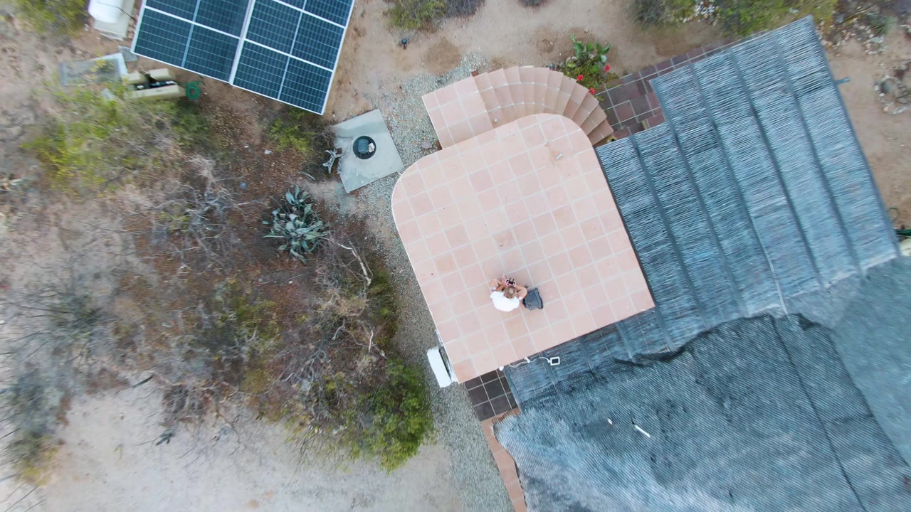 Top-down view of a desert home roof, patio, solar panels, and dry vegetation.