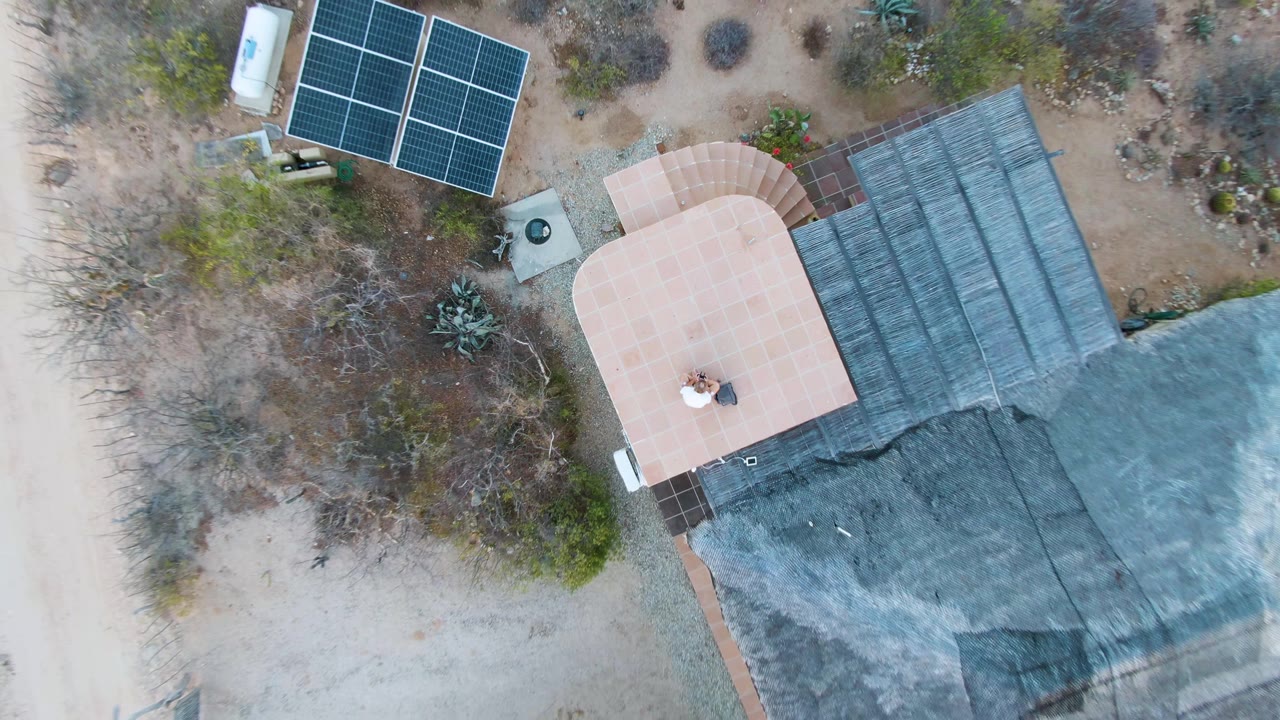 Overhead frame in Cabo Pulmo, Baja California Sur showing the house and shaded roof beside a pale dirt road, with solar panels and scattered desert plants around the property.