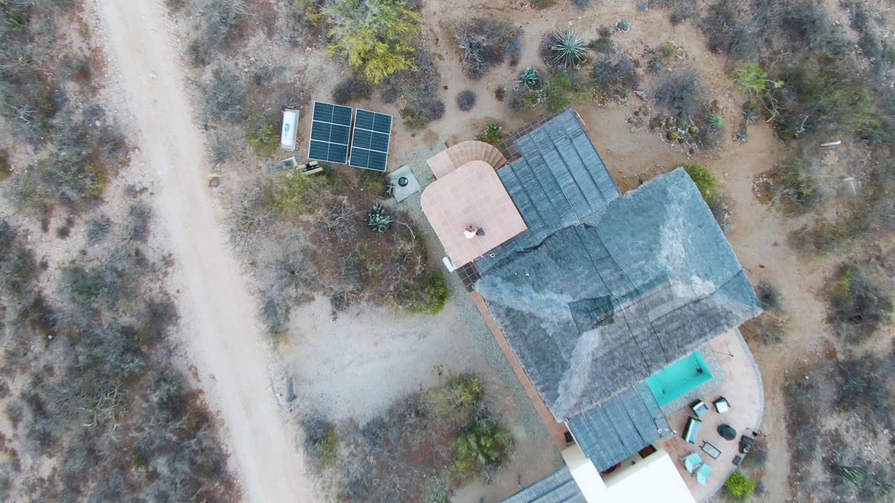 Overhead drone frame of a house in Cabo Pulmo, Baja California Sur with solar panels, a small turquoise pool, dry brush, pale dirt roads, and no visible people.
